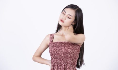 肩がこっていそうに首を傾けている黒髪のワンピース着た女性