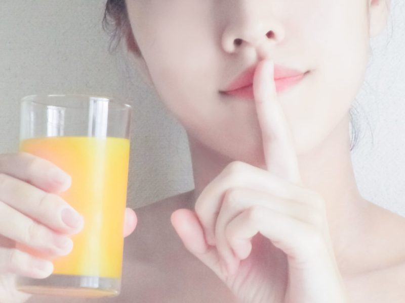 オレンジジュースを持ち口に指を当てる女性