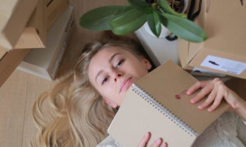 床に物がいっぱい置かれている中にノートを広げながら床に寝転ぶ女の子