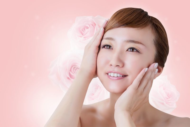 斜め上向きでデコルテから上が肌かの女性が両頬に手を添えている。背景にはピンク色をベースに同系色のバラが飾れている