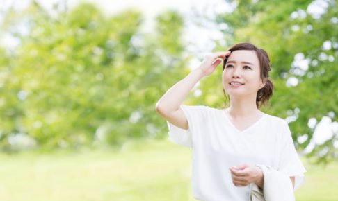 木々が生い茂る公園のなかで白色のカットソーを着た女性が額に手を当てている