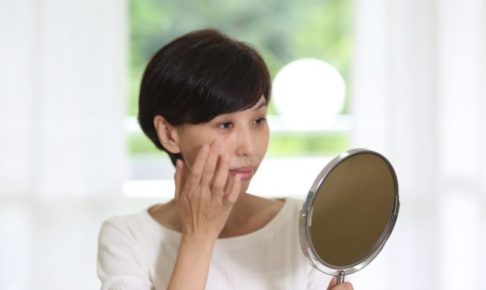 カーテンの近くで自分のしわの様子を鏡で見ているご婦人